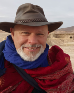 [photo: Headshot of Muiz with hat and red shawl, in the Moroccan desert, 2017